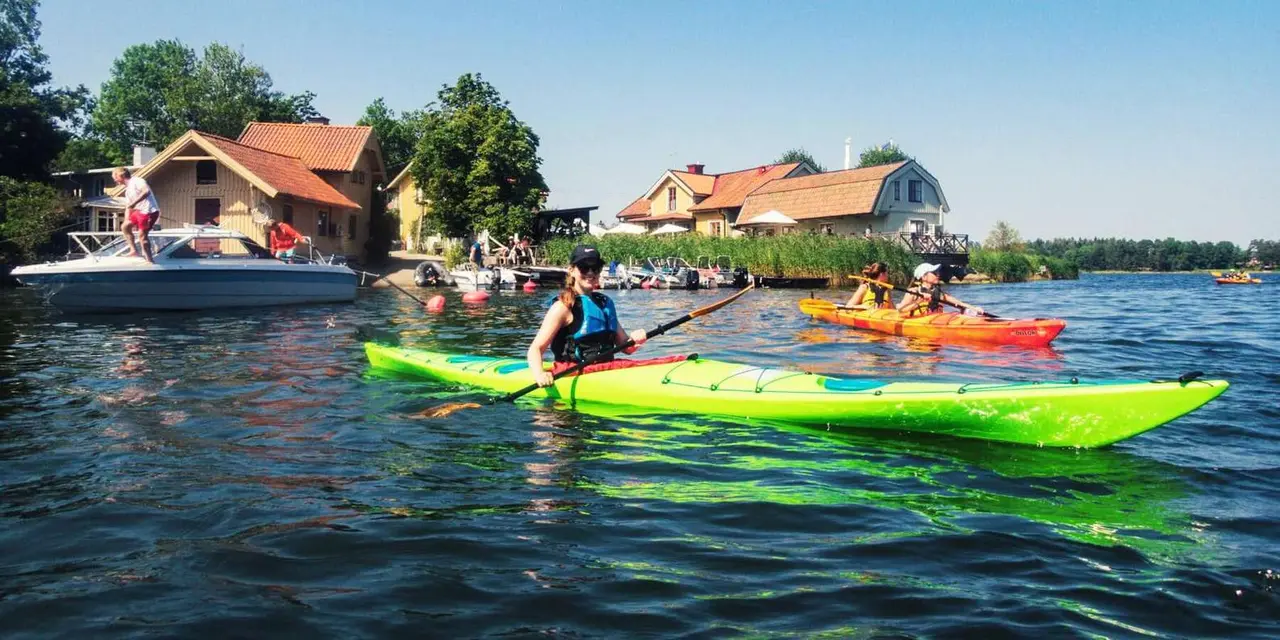A kayaker in a lime-green kayak on a calm bay, with red cottages and trees on the shore behind