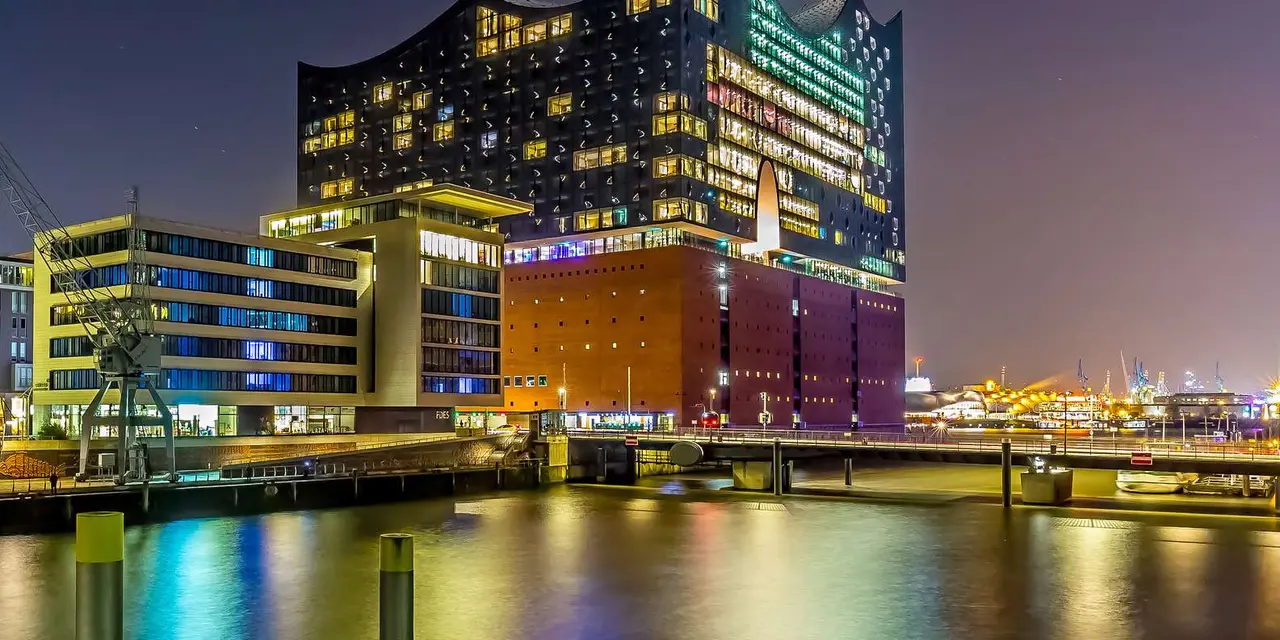 The Elbphilharmonie lit up at night above its brick base, reflected in the harbor basin, with a harbor crane in the foreground