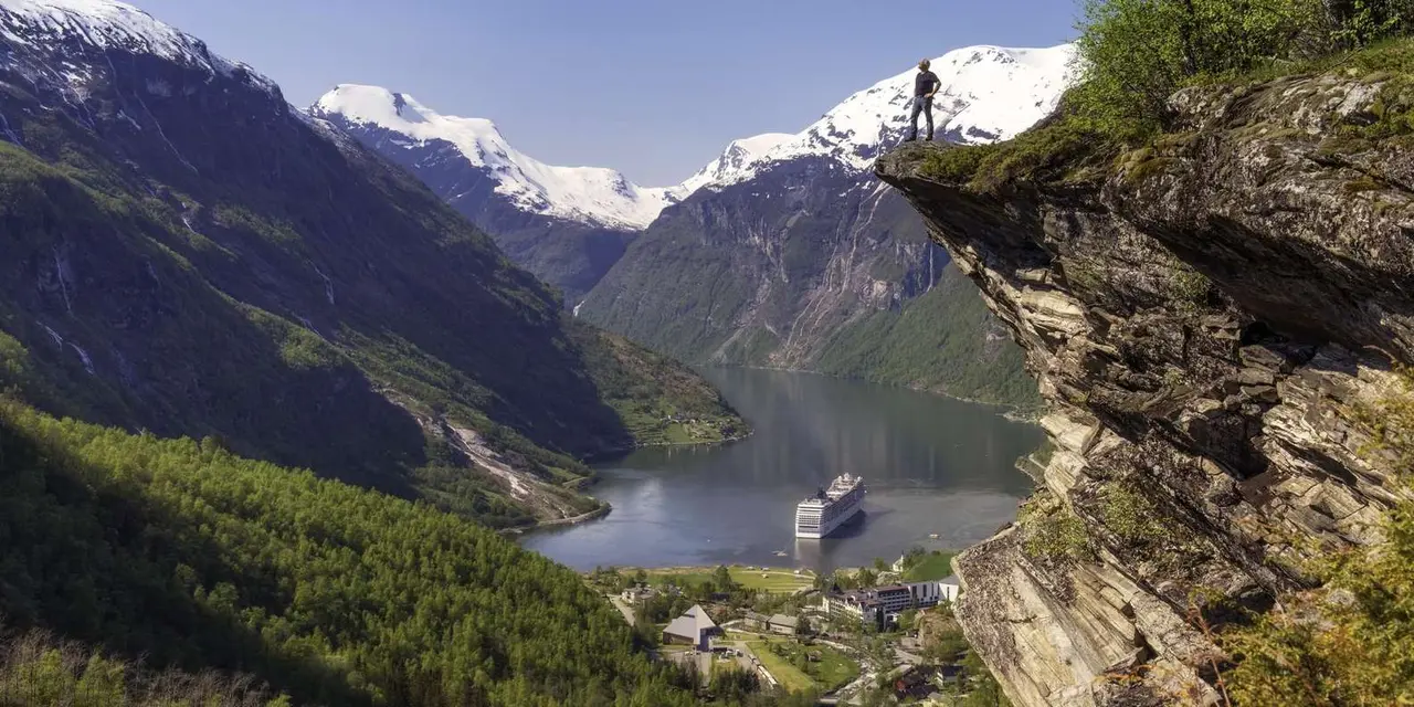 A person standing on a rocky ledge high above the Geirangerfjord, with a cruise ship far below