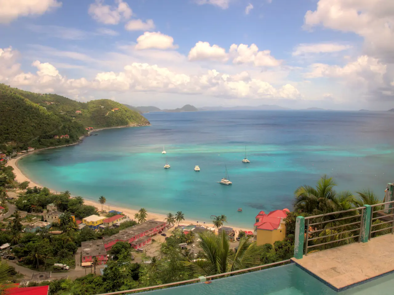 Cane Garden Bay on Tortola seen from a hillside: a curve of sand, turquoise water with sailboats at anchor, green hills and other islands on the horizon under a blue sky with white clouds