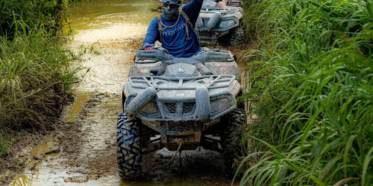Two riders on ATVs following a guide down a muddy trail between tall grass