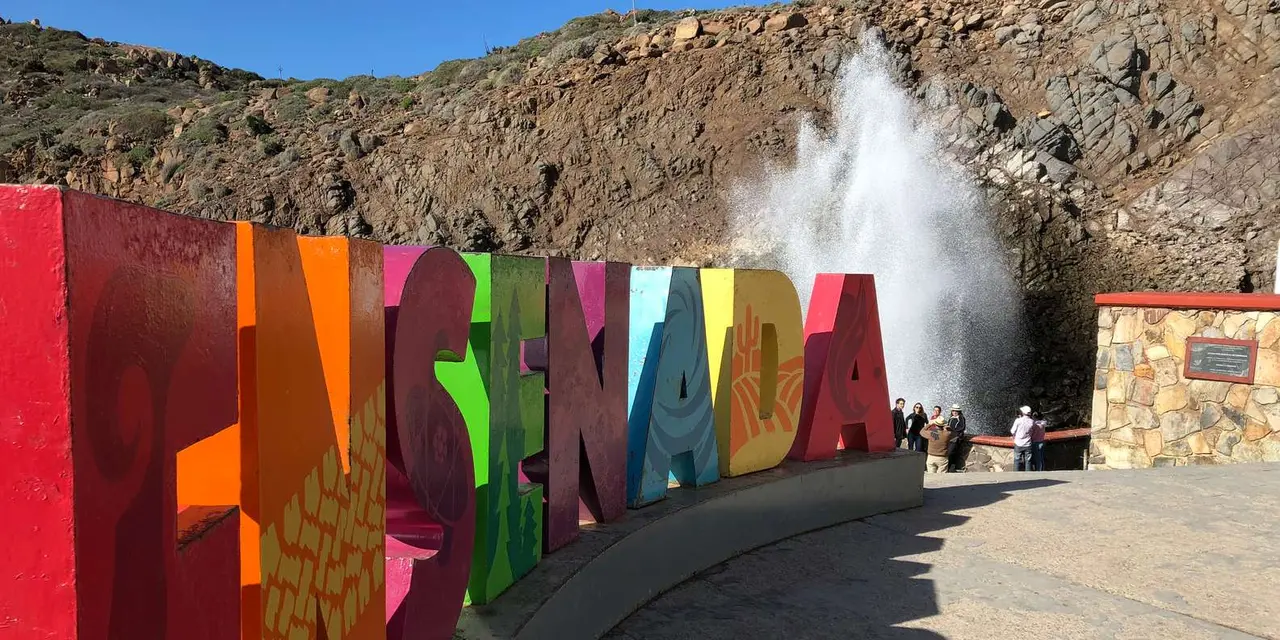 A jet of white sea spray shooting up from rocks behind a colorful letter sign, with visitors on the walkway