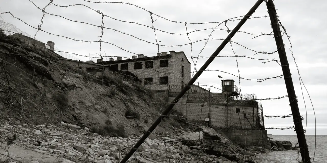 Black-and-white view through a barbed-wire fence to a weathered brick prison block with a guard tower beside the sea