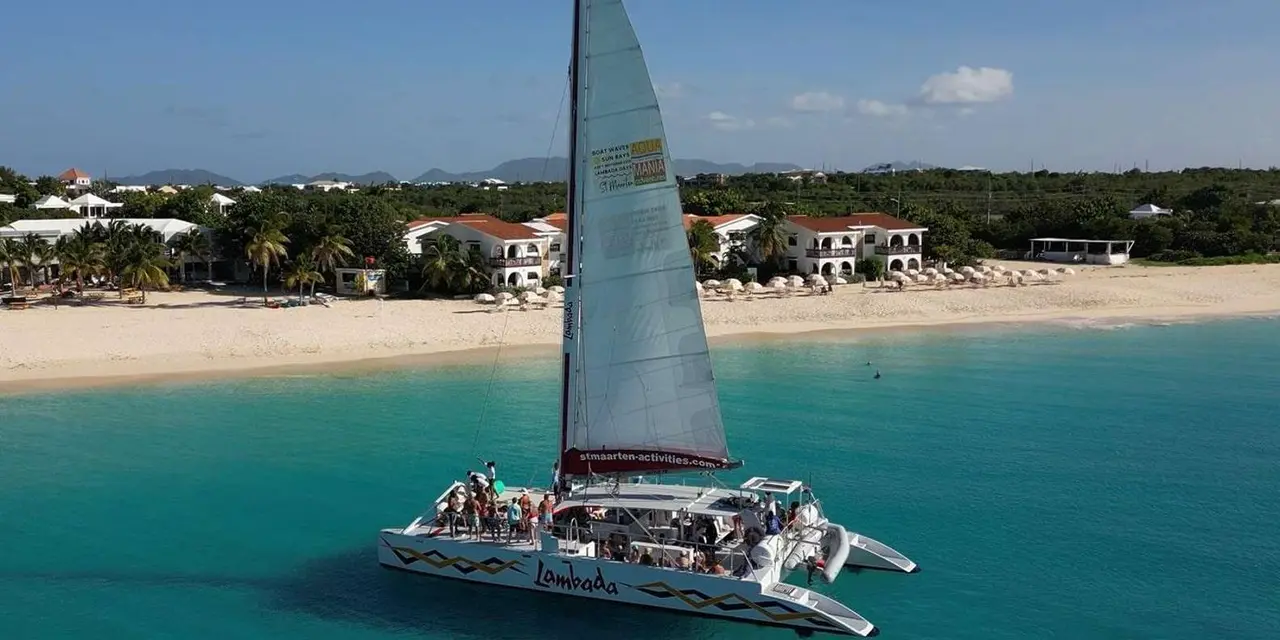 A white catamaran under sail off a long white beach lined with villas, in turquoise water