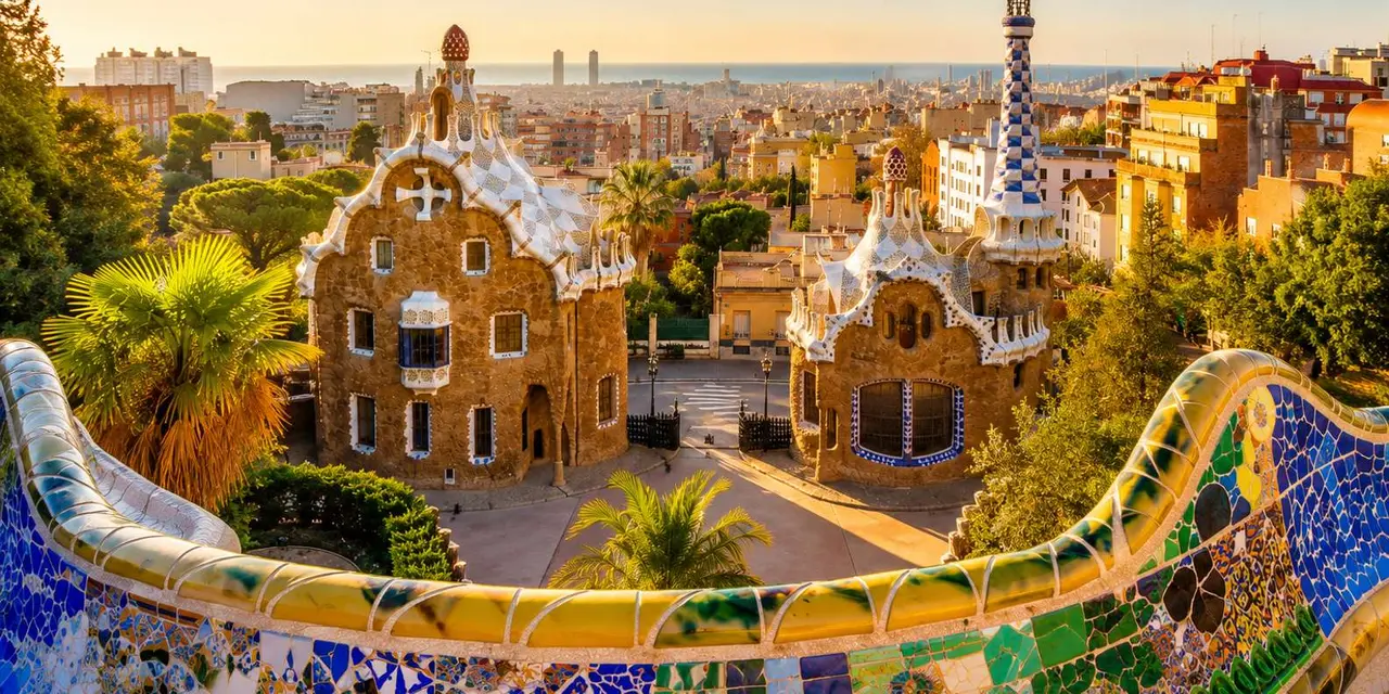 Park Güell at golden hour, two gingerbread-style gatehouses with tiled roofs and a mosaic bench in the foreground, with Barcelona stretching to the sea behind