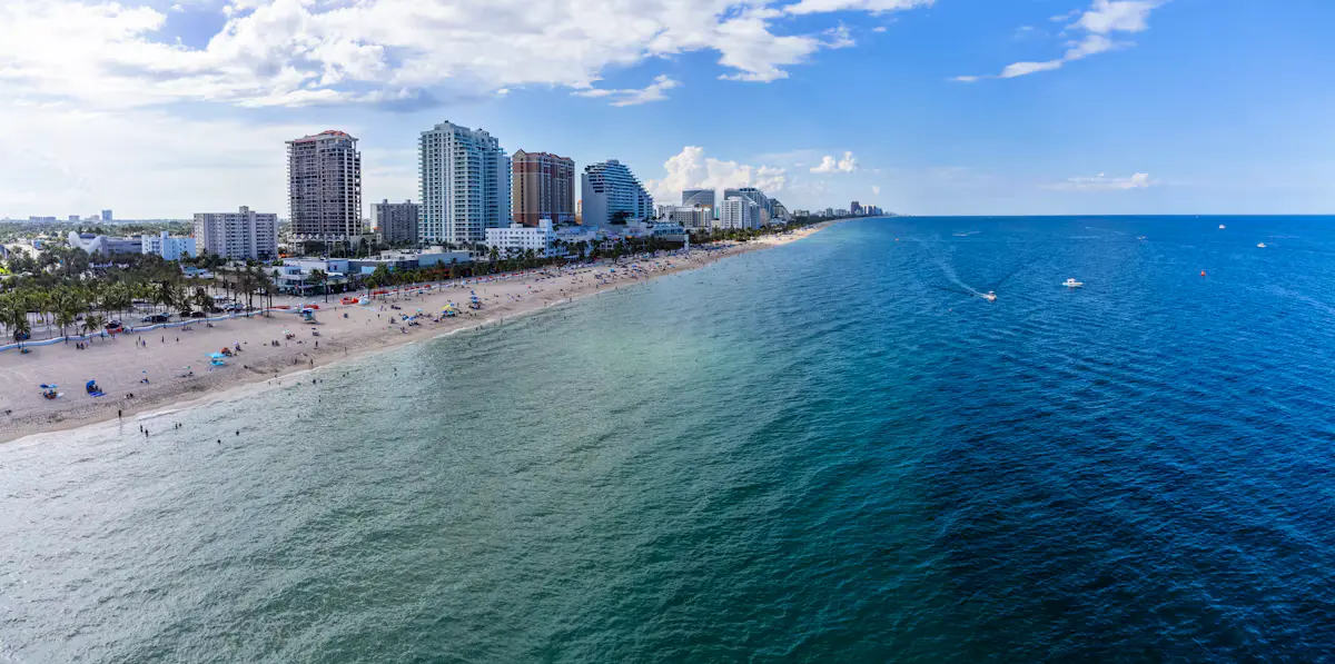 Fort Lauderdale beach seen from above: a long curve of sand lined with palms and condo towers, with turquoise shallows fading to deep blue water