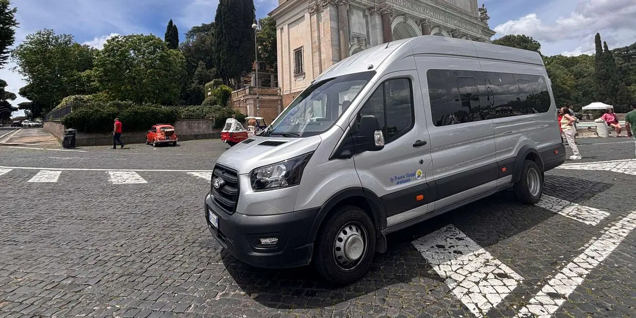 A silver tour minibus parked on a cobbled square, with a pale stone church front and tall cypress trees behind it