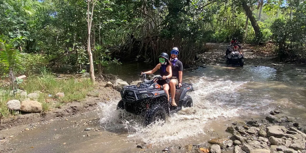 Two riders on an ATV splashing through a shallow river, with another ATV following behind