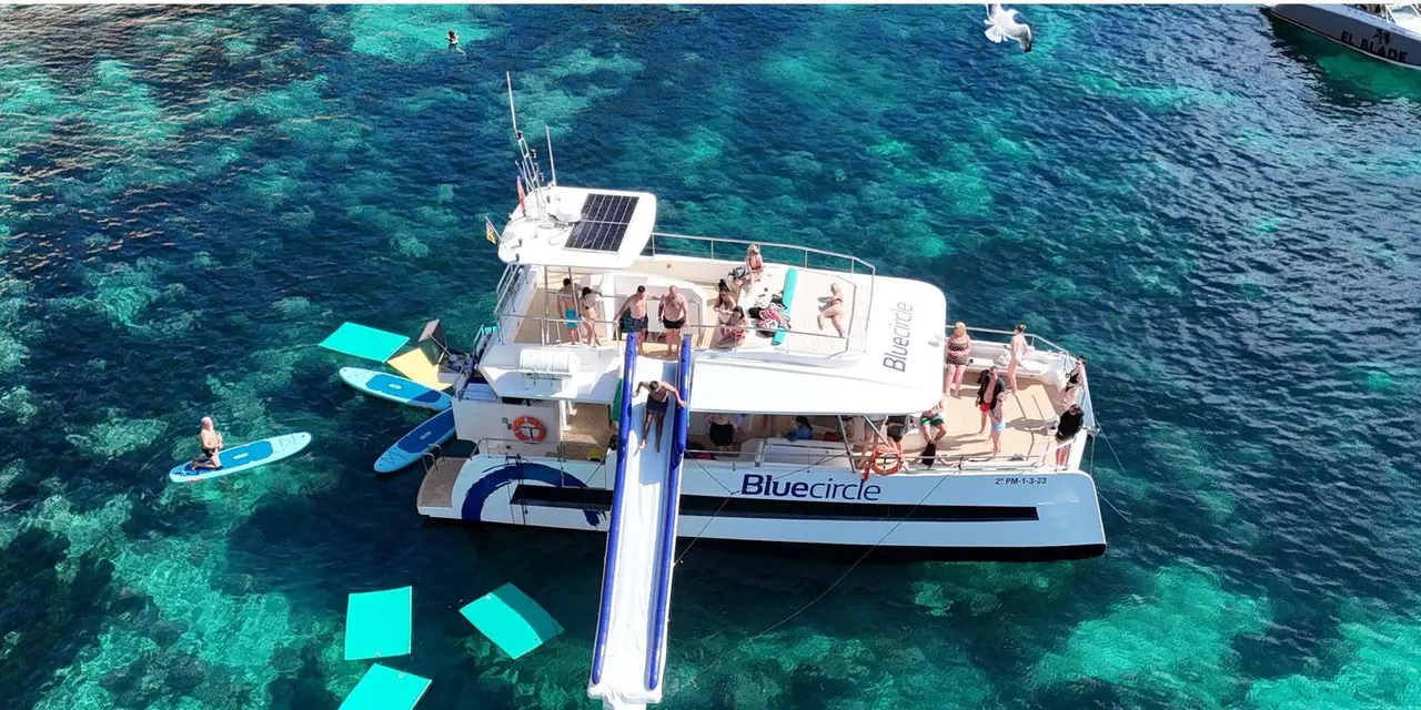 Aerial view of a white catamaran with a water slide and paddleboards floating on turquoise water