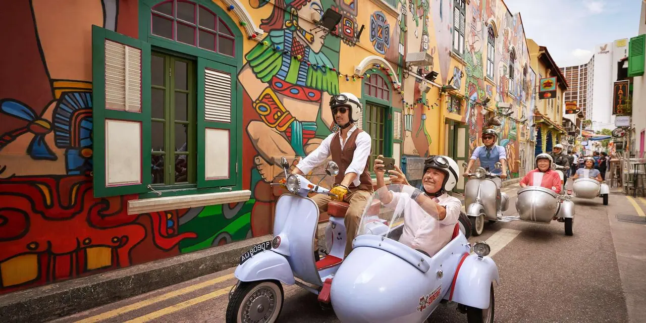 Riders in helmets in white Vespa sidecars passing a mural-covered shophouse street