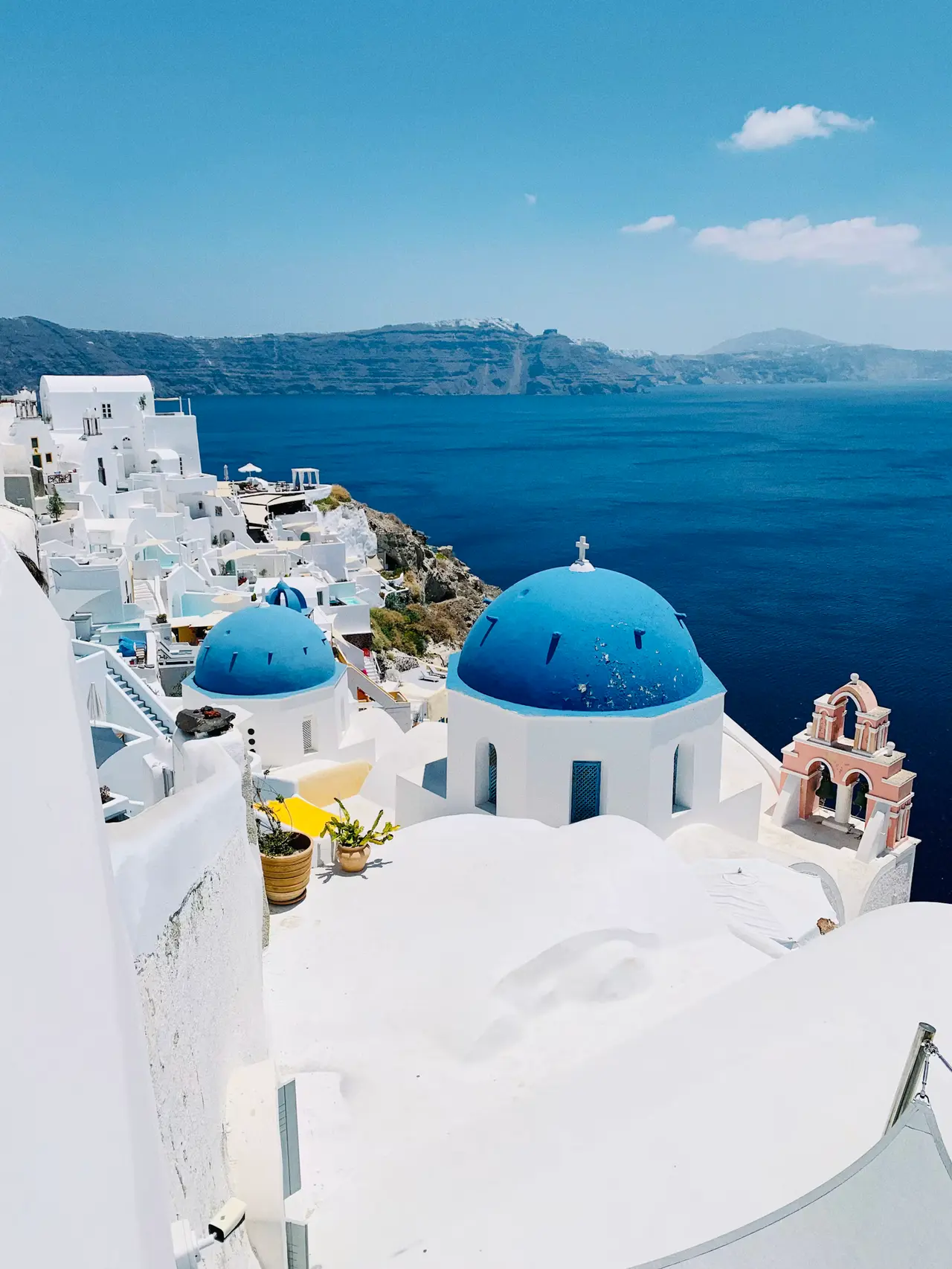Two blue-domed white churches and a pink bell tower on a Santorini cliff, with the deep blue caldera and distant rim under a clear sky