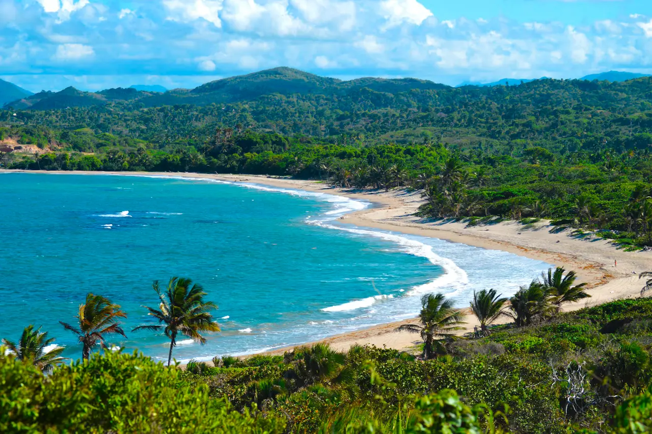 A curved bay on the Puerto Plata coast, with turquoise water, a line of white surf along a pale sand beach, palm trees and green mountains under a blue sky