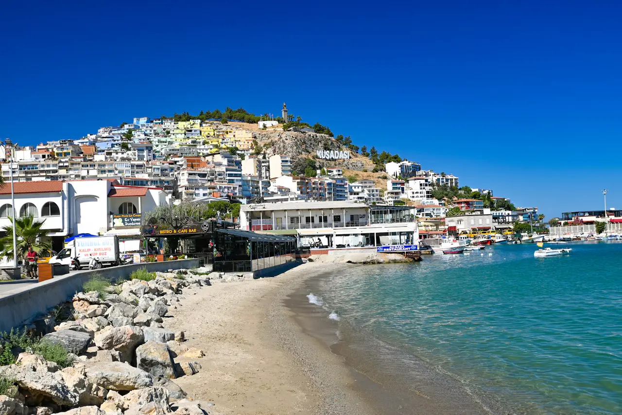 Kuşadası's town beach curving toward the harbour, with white buildings climbing the hill behind under a deep blue sky