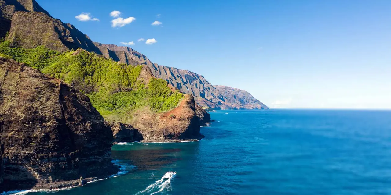 Steep green Na Pali cliffs dropping into deep blue sea, with a boat's white wake far below