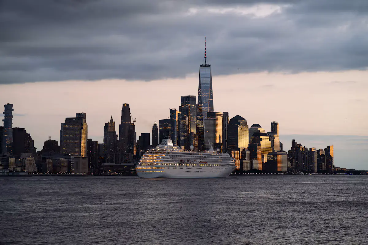 A white cruise ship with its lights on sailing the Hudson River at dusk, in front of the Lower Manhattan skyline and One World Trade Center