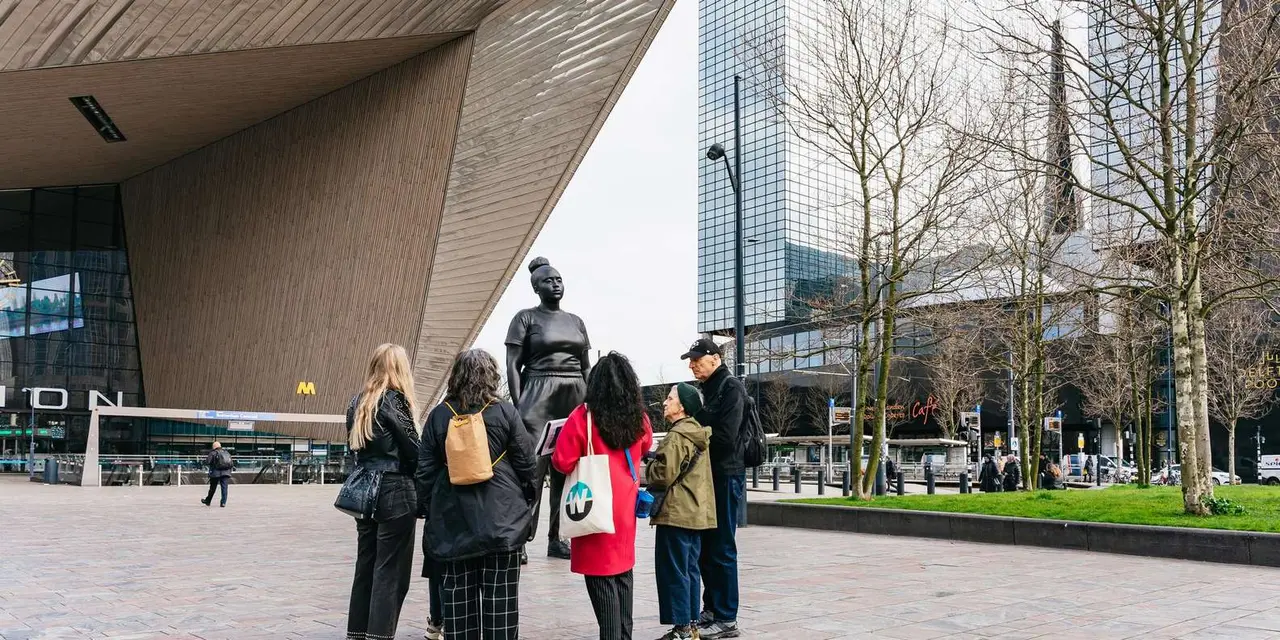 A guide and a group of six visitors stand in front of a large dark statue of a standing woman outside Rotterdam Centraal station