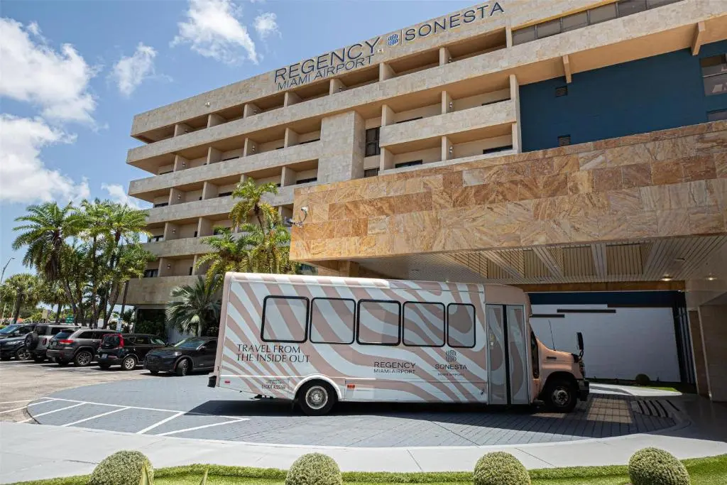 The Regency Miami Airport by Sonesta building with its striped hotel shuttle bus parked at the entrance under palm trees