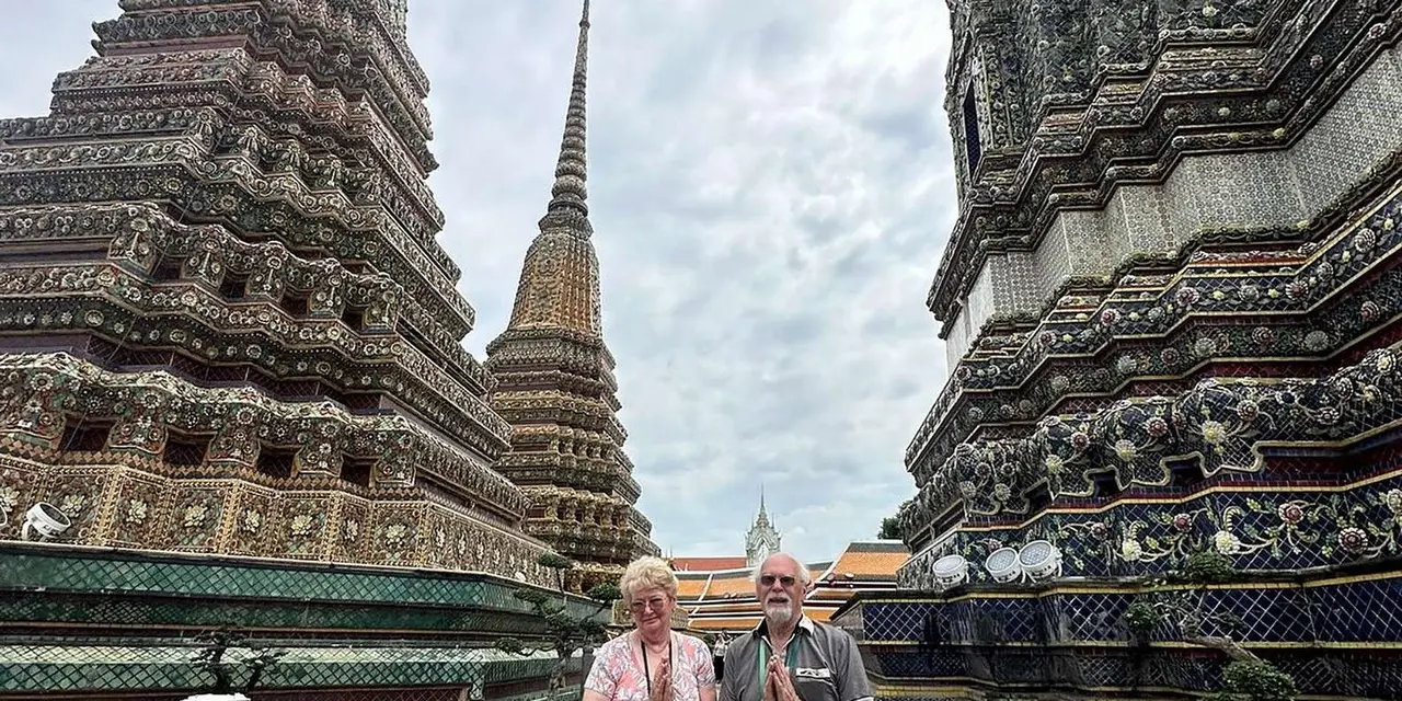 Two visitors at the foot of Wat Pho's tall tiled towers under a cloudy sky