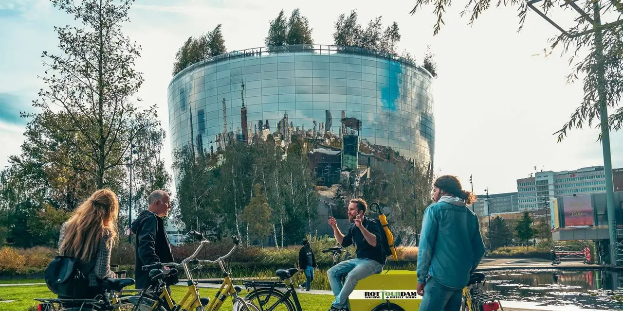 Four visitors with yellow bikes listen to a guide sitting on a cargo bike in front of the mirrored, tree-topped Depot Boijmans building