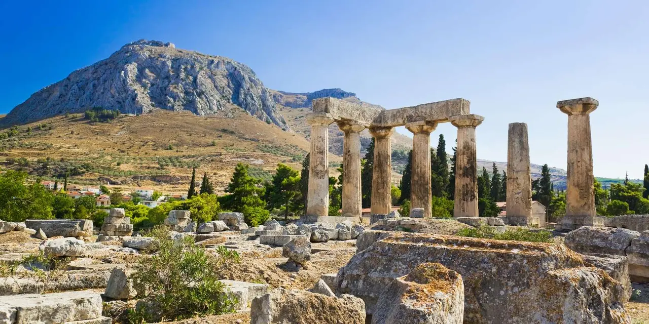 A row of weathered stone columns among ruins and pine trees, a rocky mountain rising behind