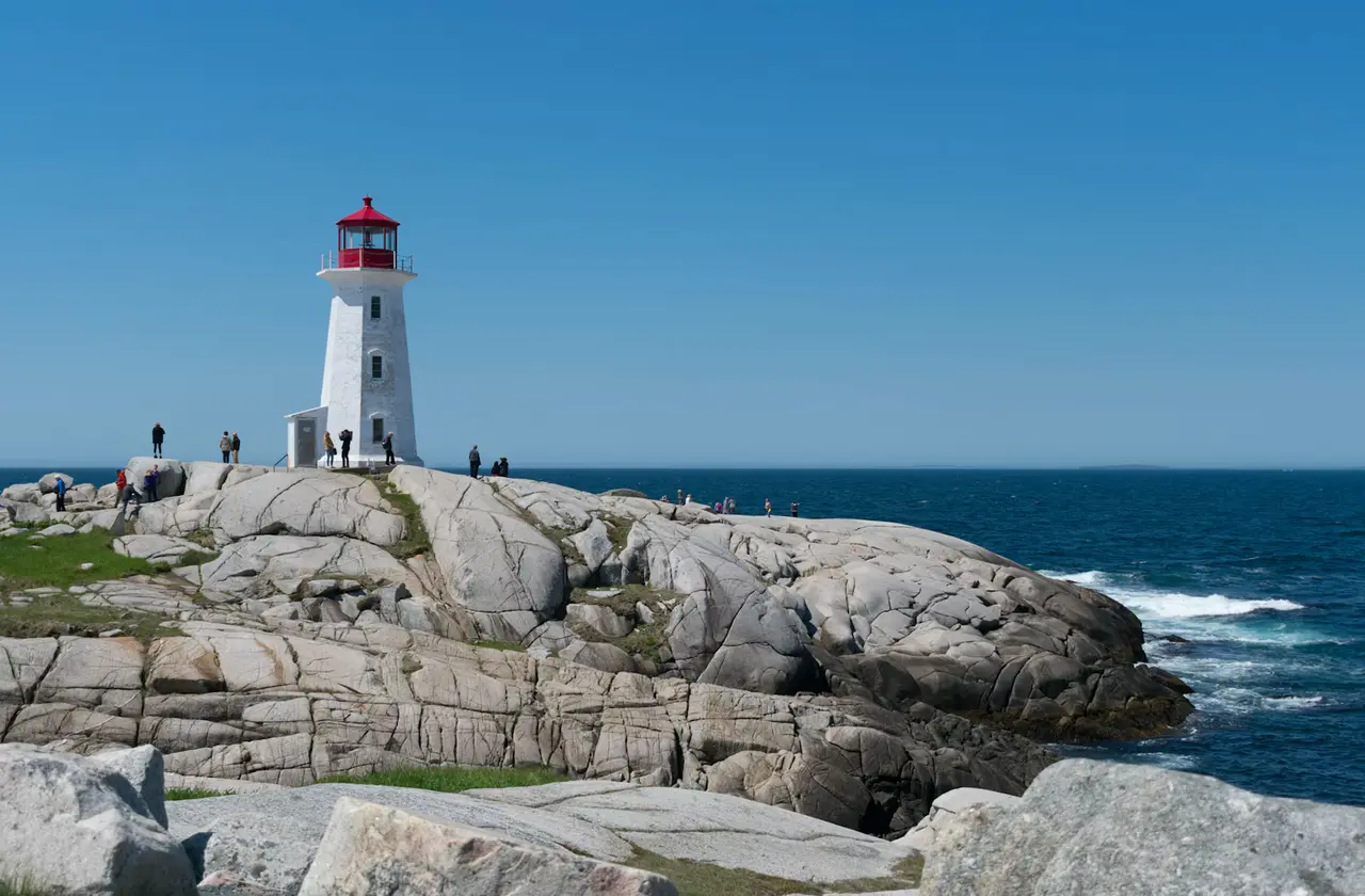 The white-and-red Peggy's Point Lighthouse on smooth granite rocks above a deep-blue Atlantic under a clear sky, at Peggy's Cove, about an hour's drive from the Halifax cruise port