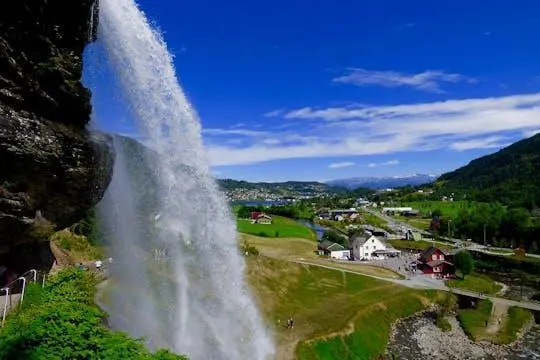 A tall waterfall pouring off a cliff into a green valley dotted with farmhouses under a blue sky