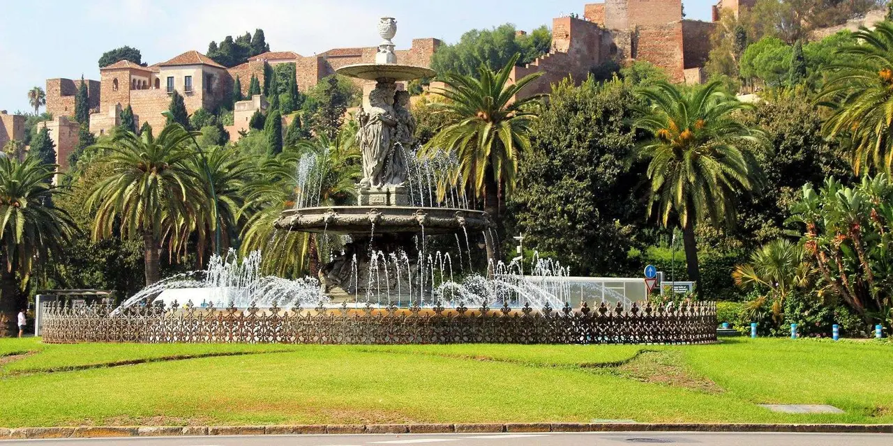 A fountain among palm trees in a park, with old buildings on the hillside above
