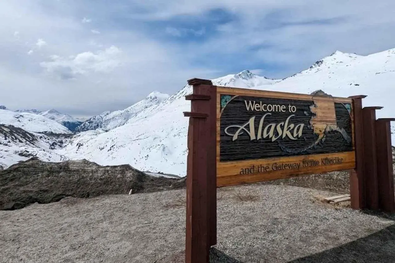 A carved wooden Welcome to Alaska sign beside snow-covered mountains