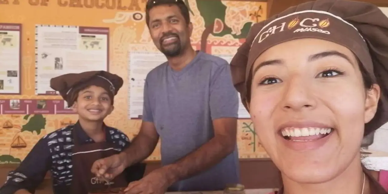 A man and a boy in brown chocolate-workshop caps and aprons smiling beside a woman in a matching cap in front of a chocolate-history display