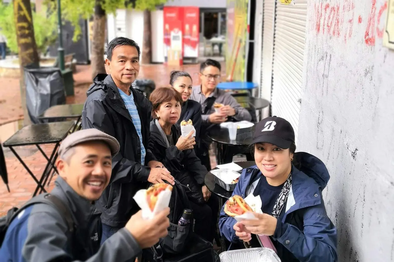 A group of smiling travelers sitting at a street-side table eating pita wraps