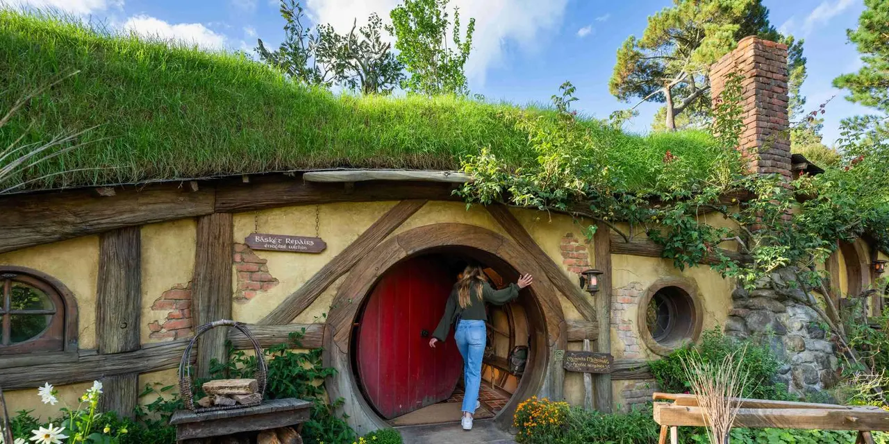 A visitor stepping up to a round red hobbit-hole door set into a grassy bank, with a garden path in front