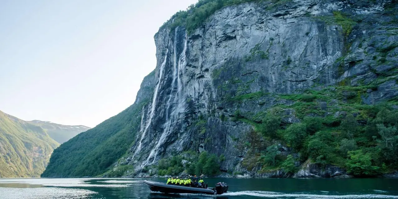 A small RIB boat at the foot of a tall cliff streaked with waterfalls