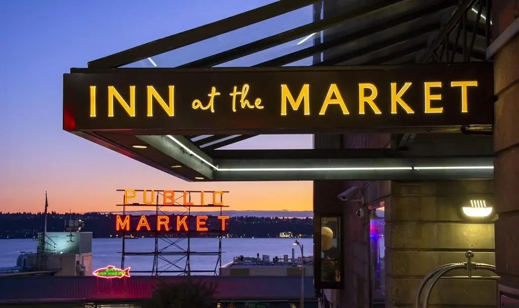The Inn at the Market sign lit at dusk with the Pike Place Market neon sign and Elliott Bay behind it