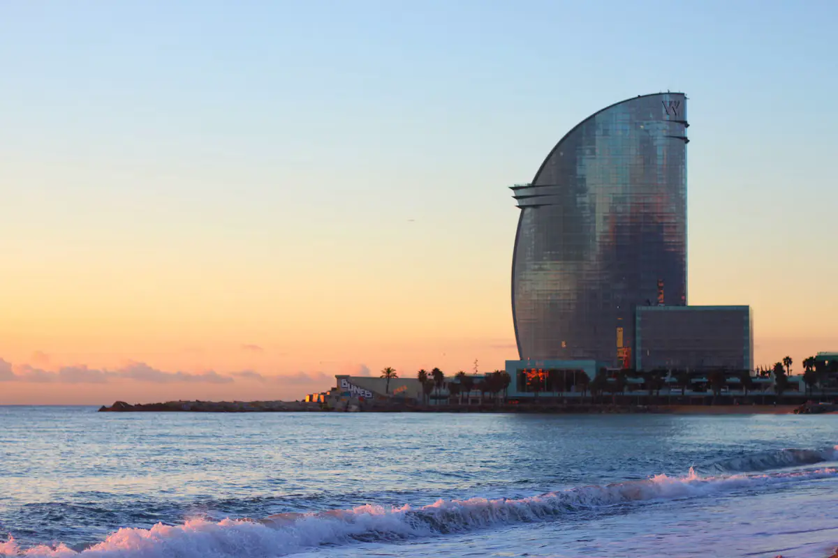 The sail-shaped W Barcelona hotel at the end of Barceloneta beach, with waves breaking on the sand under an orange sunrise sky