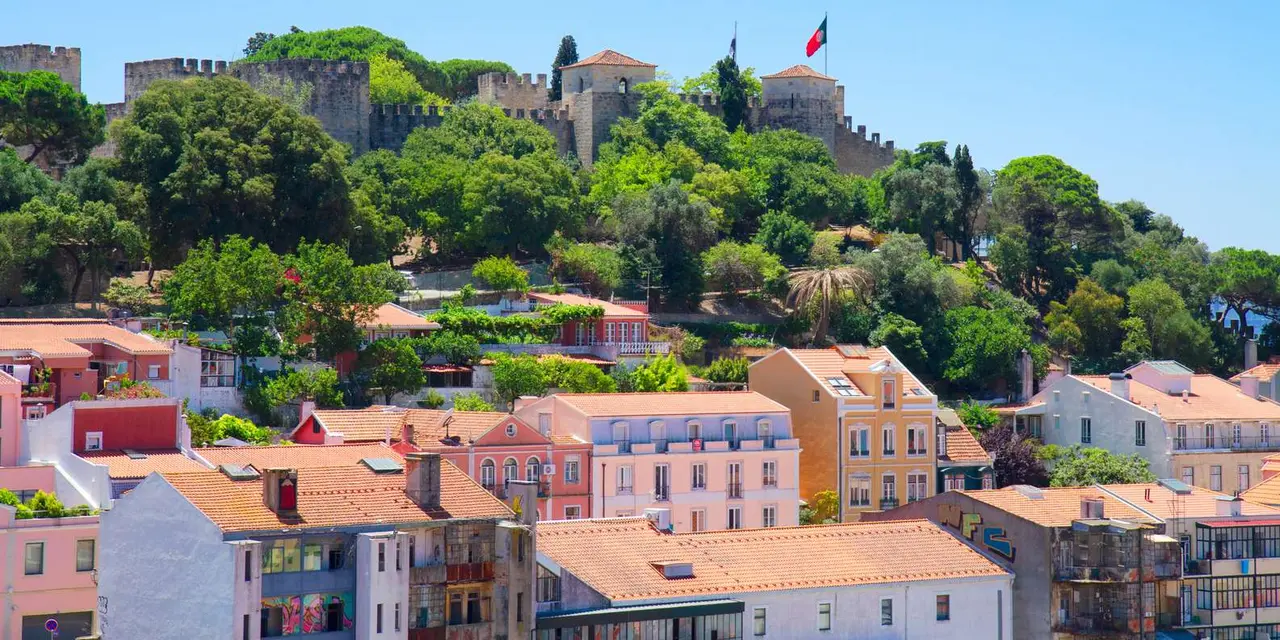 The crenellated walls and towers of São Jorge Castle with a Portuguese flag, seen across green trees and the terracotta roofs of Alfama