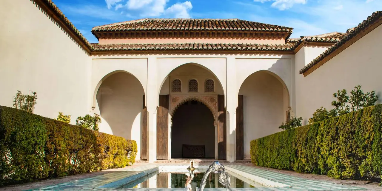 A white arcaded courtyard with arched doorways, a reflecting pool and clipped hedges inside the Alcazaba of Málaga