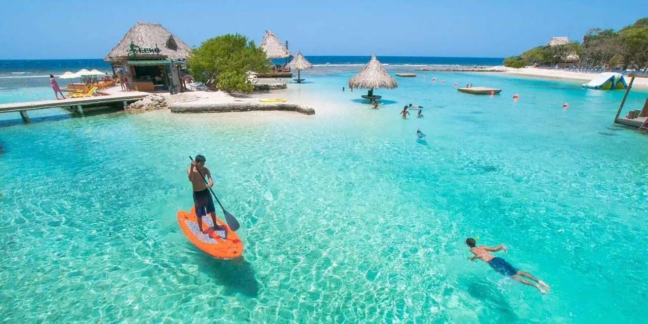 A paddleboarder and a swimmer in bright turquoise water beside a small cay with thatched huts and a tree