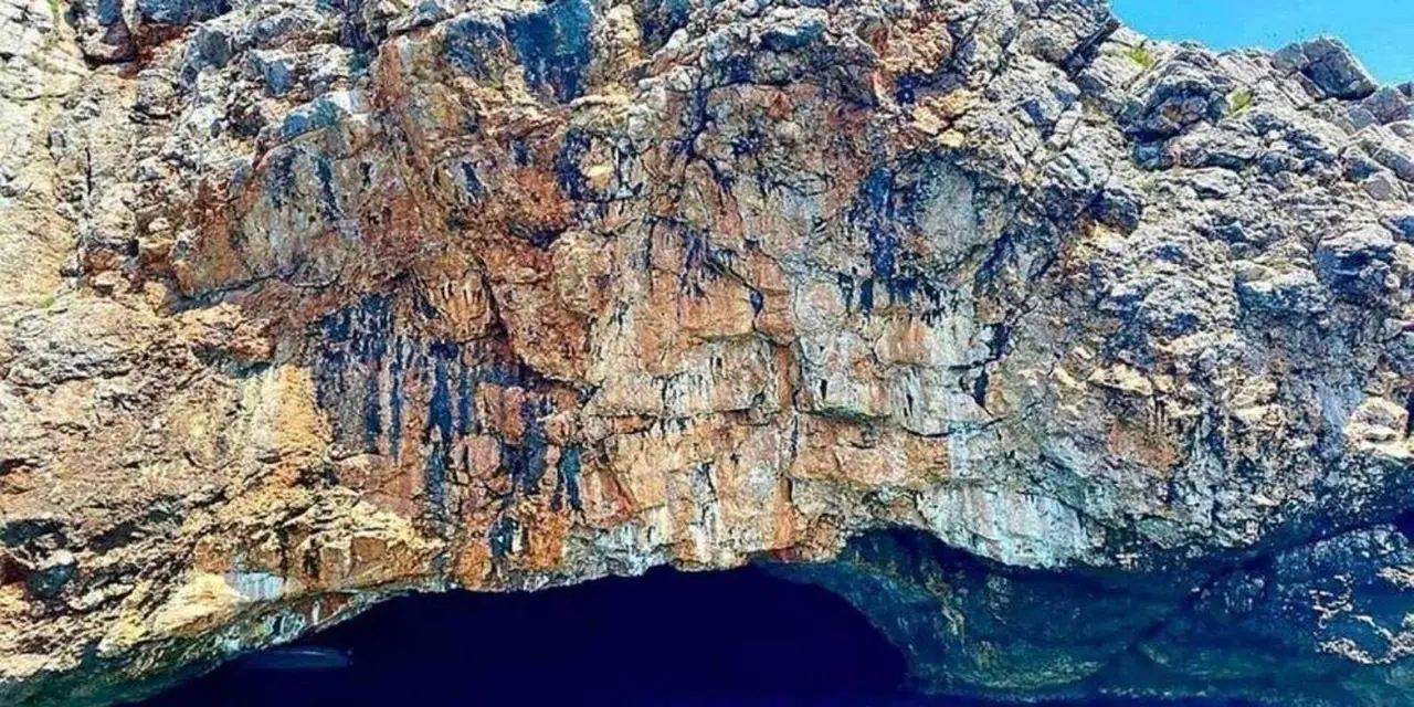 A sunlit limestone cliff with a dark cave opening at the waterline