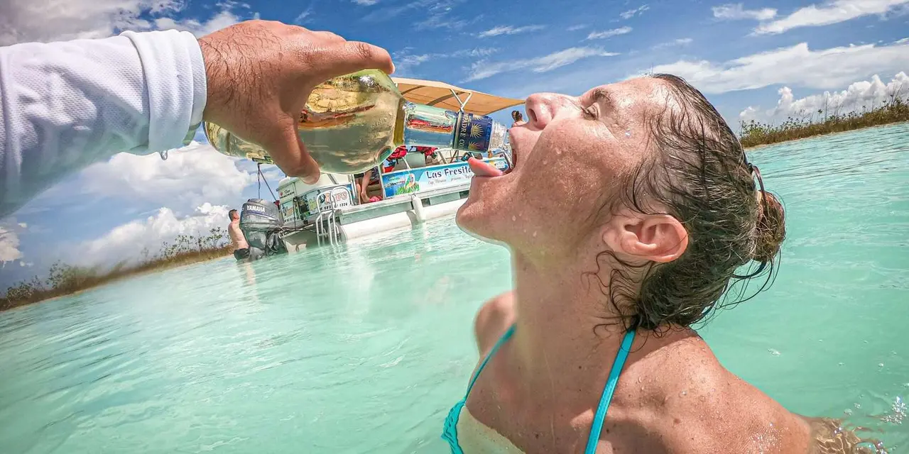 A pontoon boat with a woman at the stern pouring a drink into the mouth of a swimmer in turquoise shallows