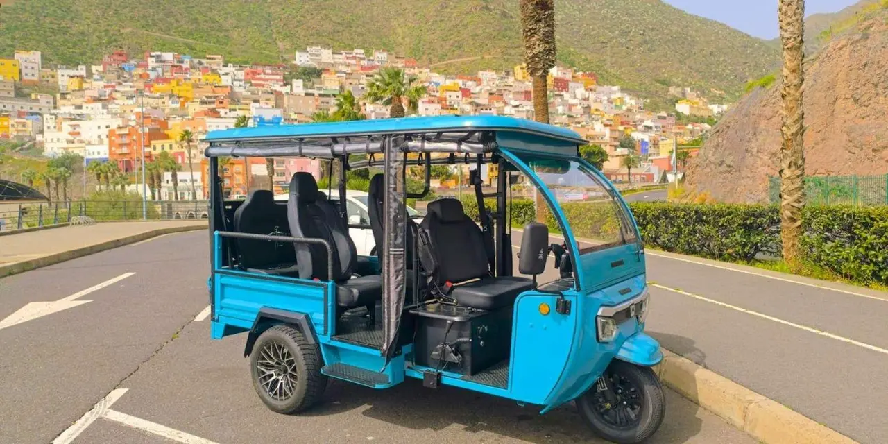 A blue open-sided tuk-tuk parked on a road with palm trees and a hillside of colourful houses behind it
