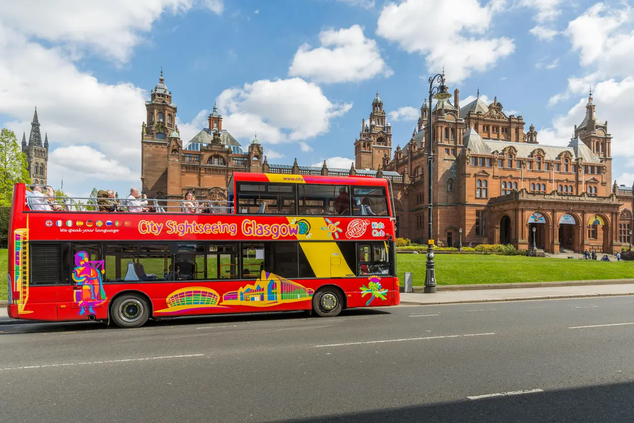A red open-top City Sightseeing Glasgow bus parked in front of Kelvingrove Art Gallery's red sandstone towers