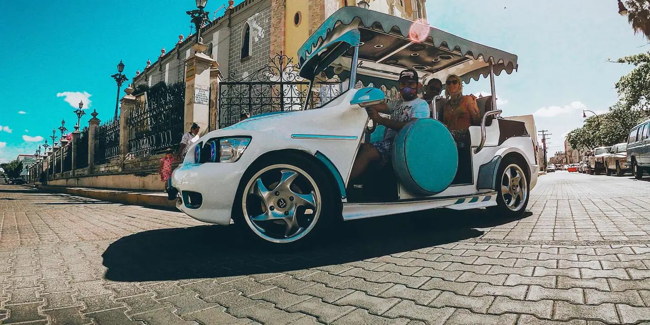 A white open-air pulmonia cab with a turquoise spare-wheel cover and passengers aboard, parked on cobblestones in front of a church