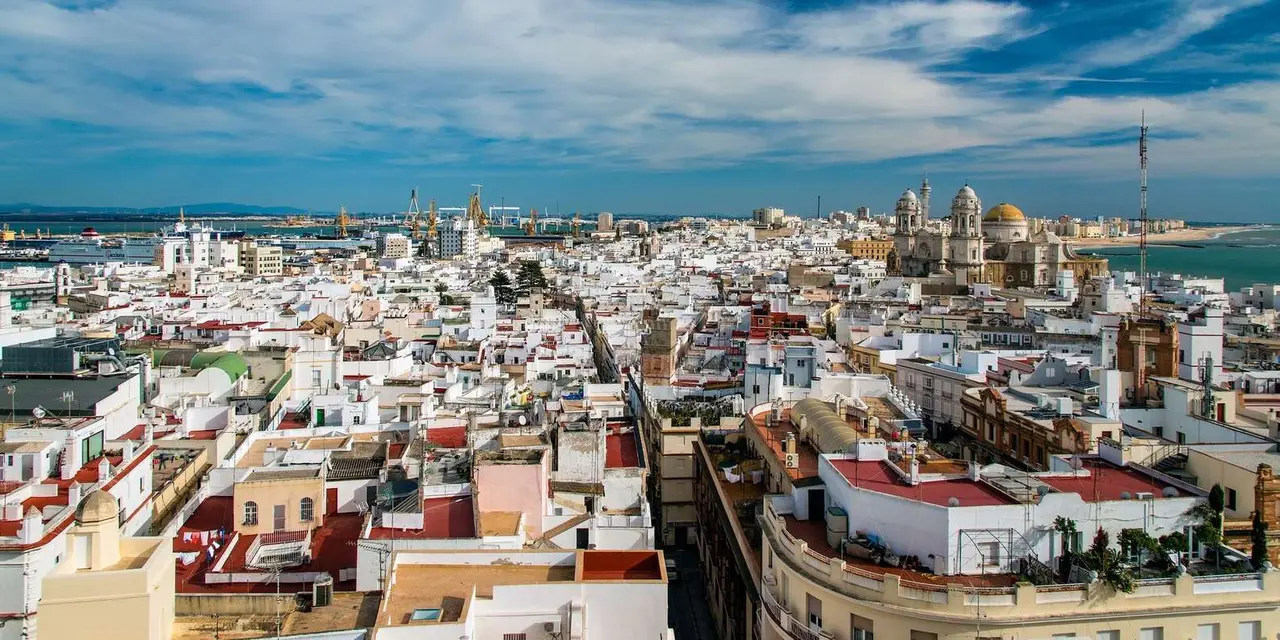 A rooftop view over Cádiz's white flat-roofed old town, with the cathedral's gold dome and towers in the distance and the sea beyond