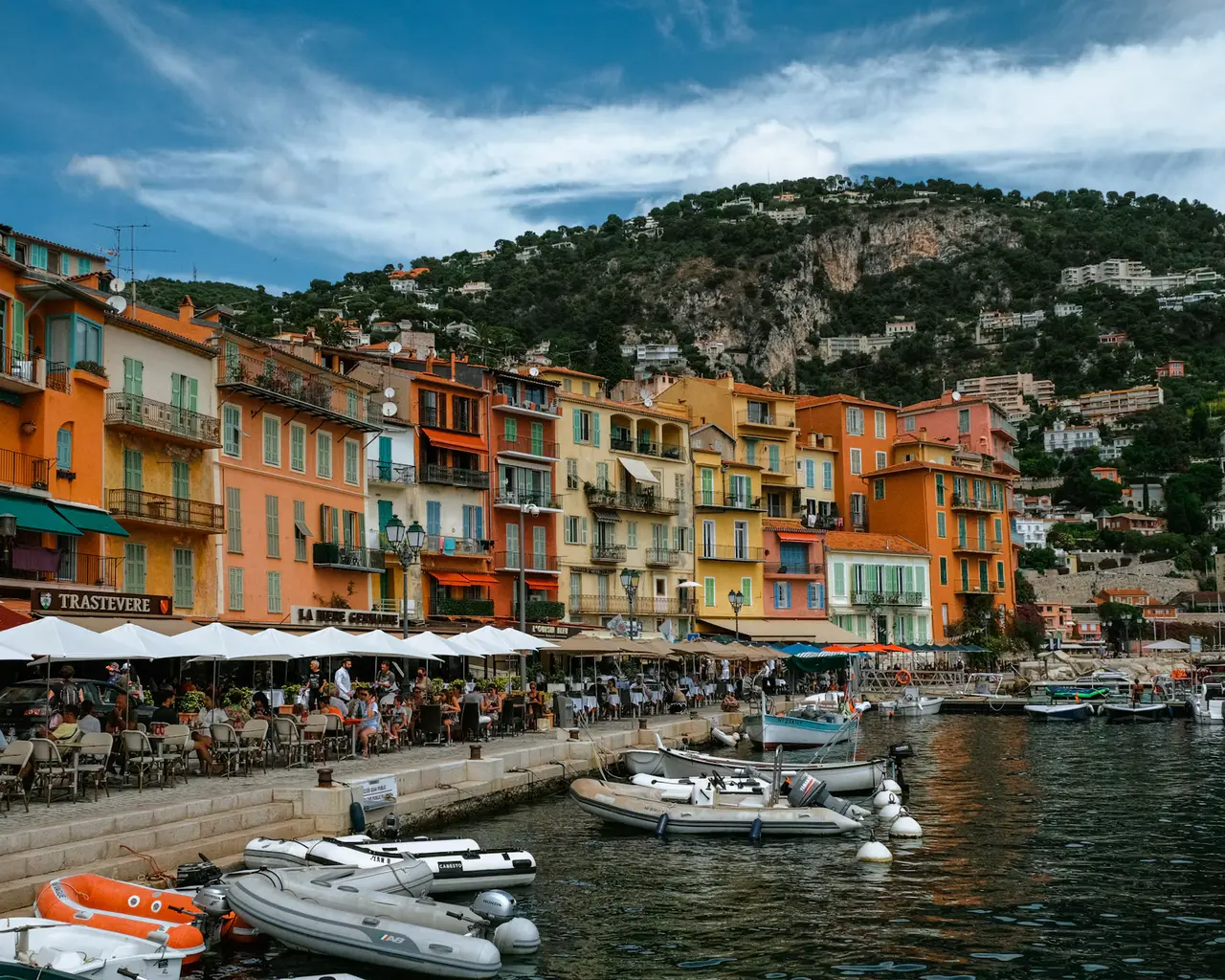 Orange and ochre houses with café awnings lining Villefranche-sur-Mer's waterfront, small boats moored in front and a green hill under a blue sky with white clouds