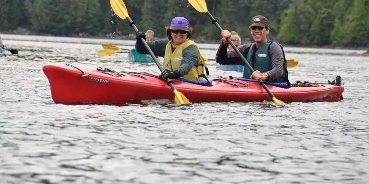Two kayakers in red kayaks on calm water with forest behind