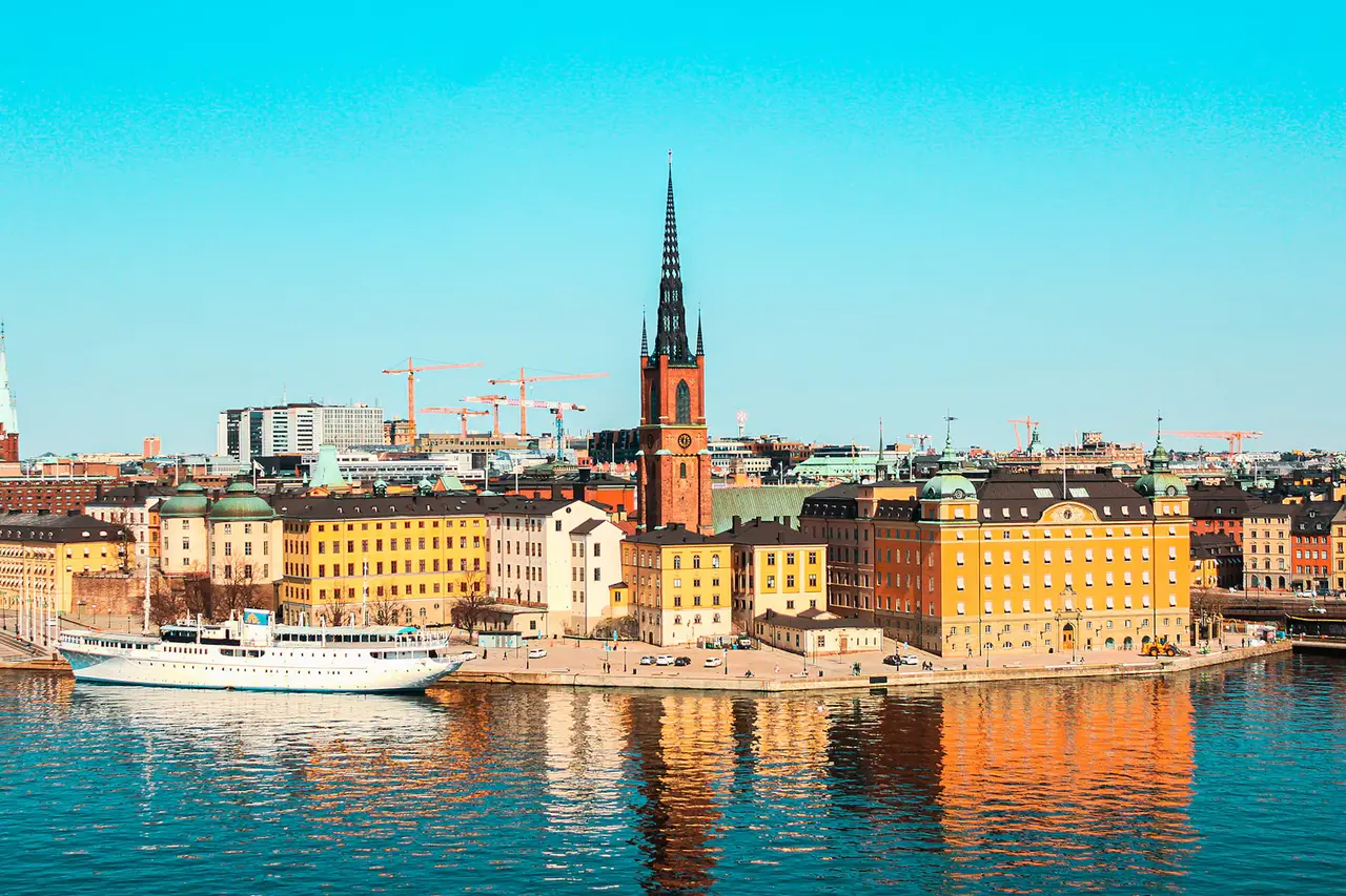 A white passenger boat on turquoise water in front of the pastel and brick waterfront buildings of Stockholm's Old Town, with a tall dark church spire rising above them under a bright blue sky