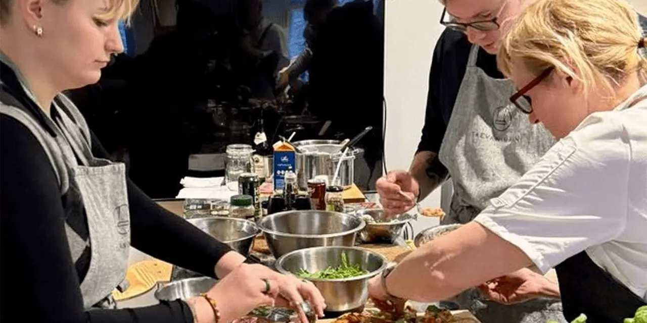 Three people in grey aprons preparing food around a table of steel bowls, with a chef in white on the right