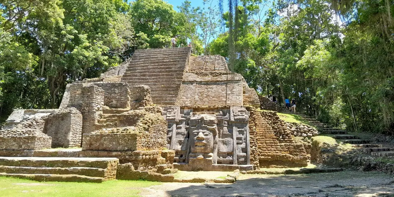 A stone Maya temple with large carved masks on its face, in a forest clearing