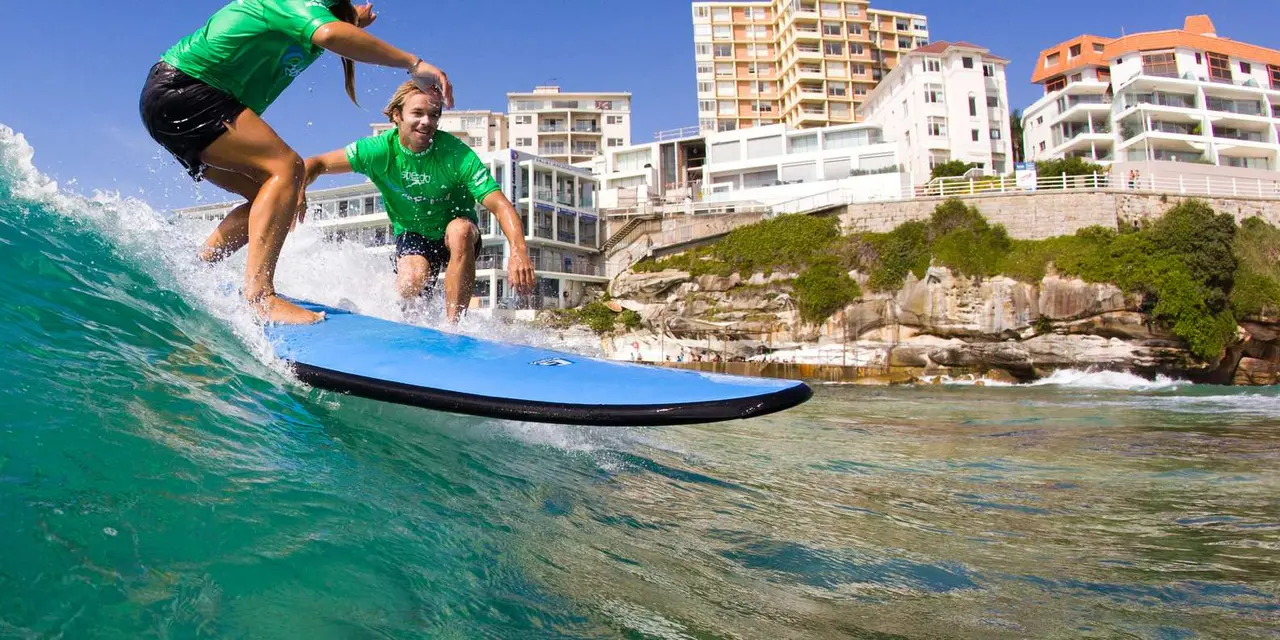 Two surfers in green shirts riding a wave at Bondi, with the headland and apartment blocks behind