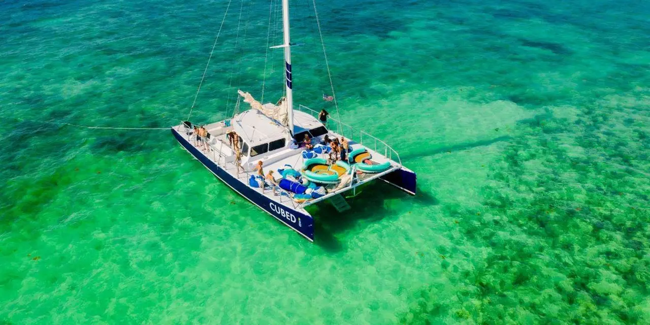 A white catamaran with a blue hull marked Cubed, seen from above in shallow green water, with passengers on its deck